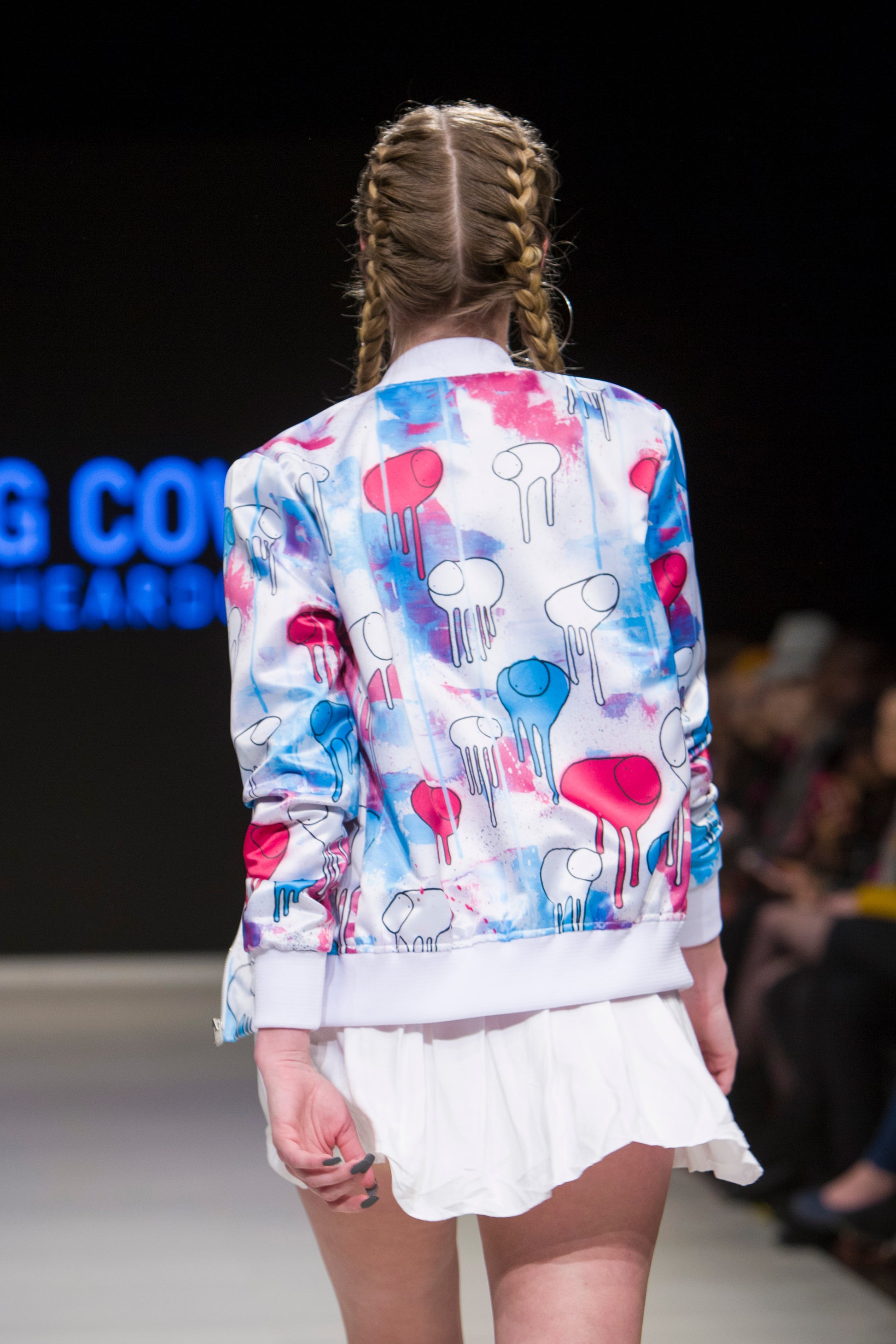 Close-up of a model turned around on the runway showing the back of a white, blue and pink multi coloured RAINING COWS bomber jacket by Morgan Sheardown, paired with white mini skirt, with seated audience members in the background.