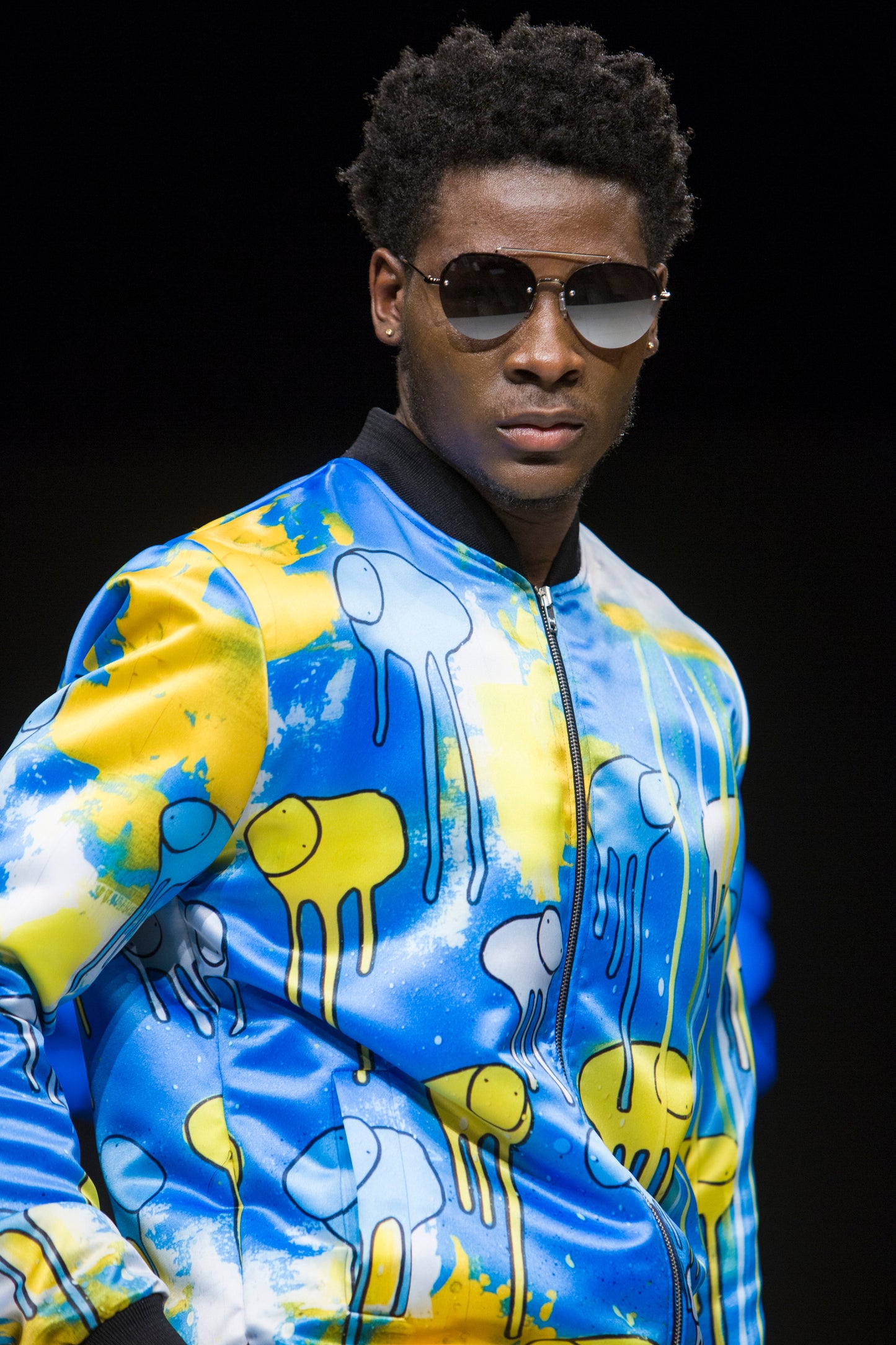 Close-up of a model turned around on the runway showing the front of a blue and yellow multi coloured RAINING COWS bomber jacket by Morgan Sheardown, zipped up, paired with sunglasses.