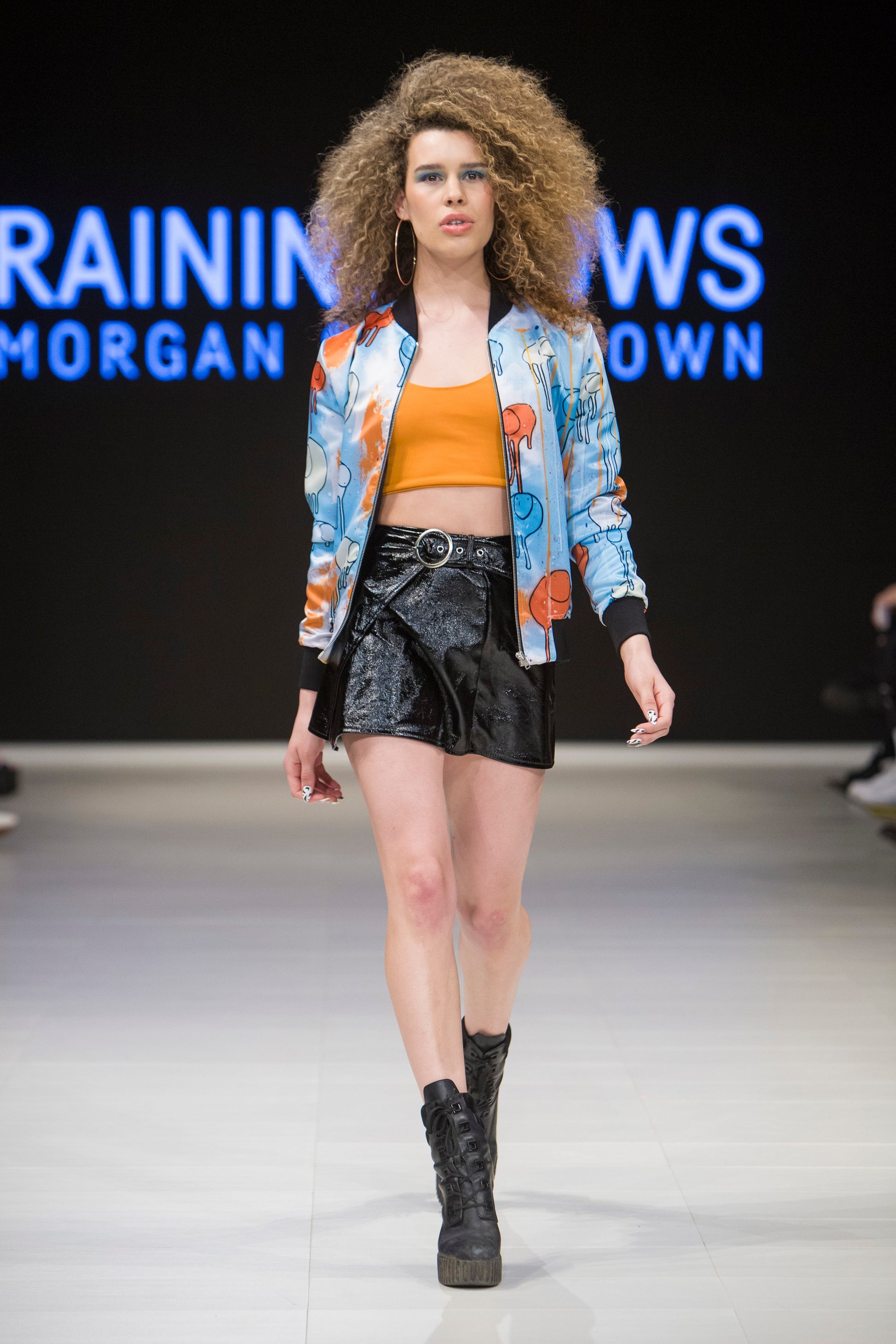 Full-body shot of a model walking the runway wearing a white, blue and orange multi coloured RAINING COWS bomber jacket by Morgan Sheardown, styled with a orange crop top, shinny black mini skirt, black high heels.