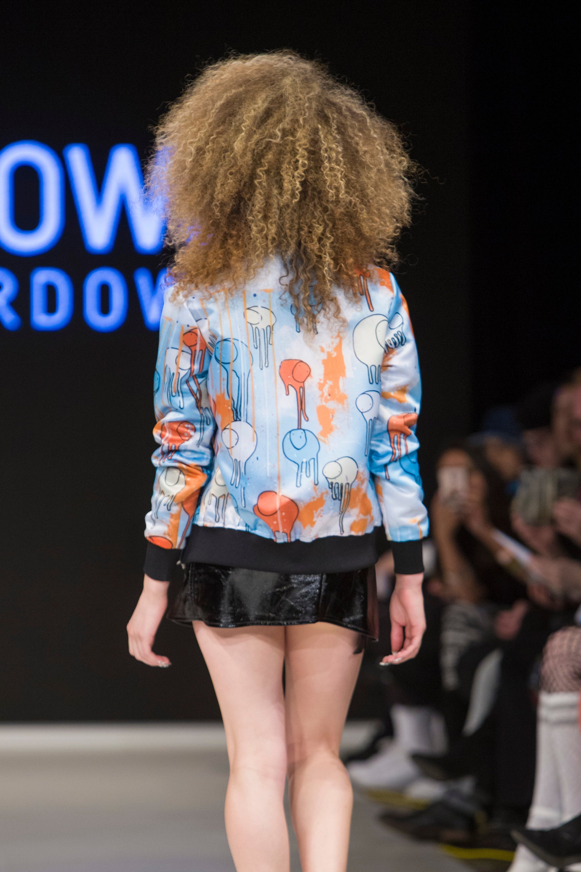 Close-up of a model turned around on the runway showing the back of a blue and orange multi coloured RAINING COWS bomber jacket by Morgan Sheardown, paired with mini skirt, with seated audience members in the background.
