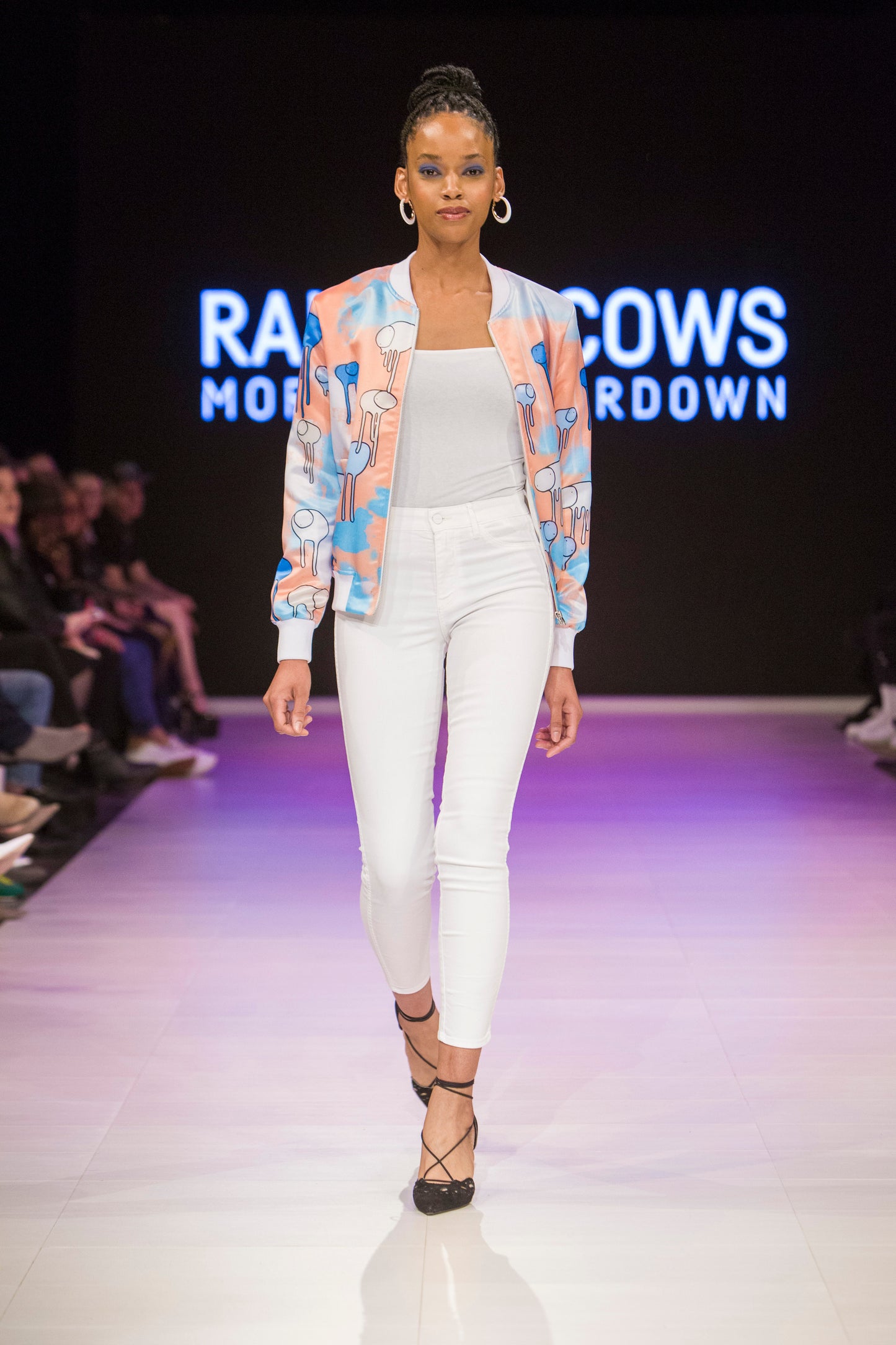 Full-body shot of a model walking the runway wearing a white, blue and orange multi coloured RAINING COWS bomber jacket by Morgan Sheardown, styled with a white crop top, white pants, black high heels.