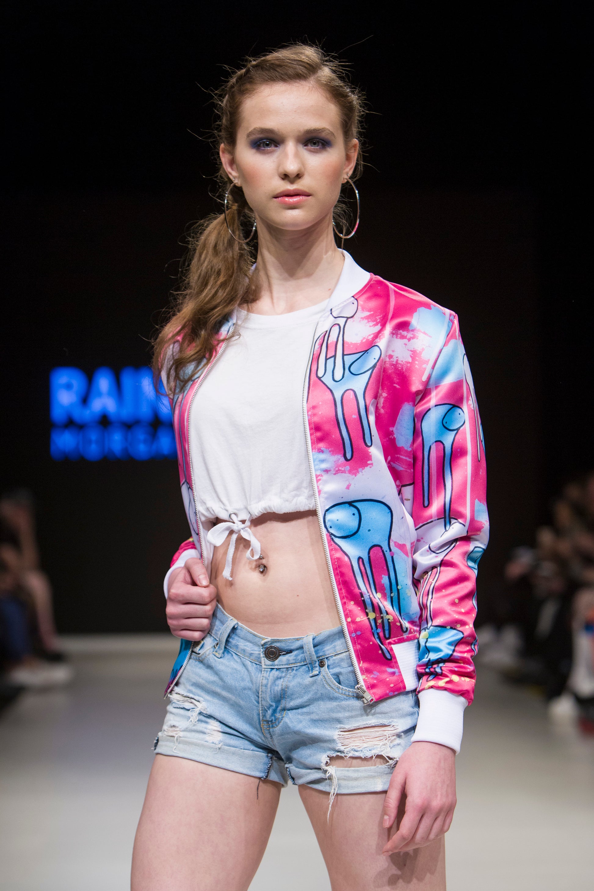 Female model wearing RAINING COWS ‘Sentiment of Colour’ bomber jacket by Morgan Sheardown, pink and blue silk with cow and rain motif, styled with white tied crop top, blue shorts, posing on the runway.
