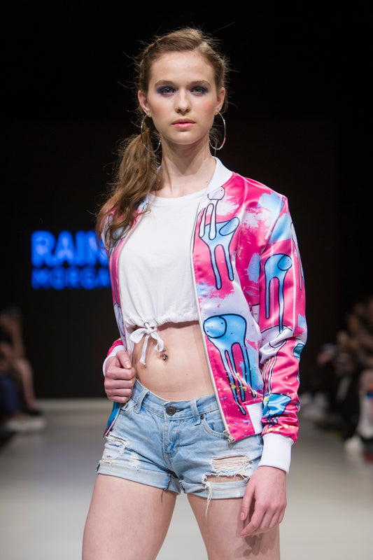Female model wearing RAINING COWS ‘Sentiment of Colour’ bomber jacket by Morgan Sheardown, pink and blue silk with cow and rain motif, styled with white tied crop top, blue shorts, posing on the runway.
