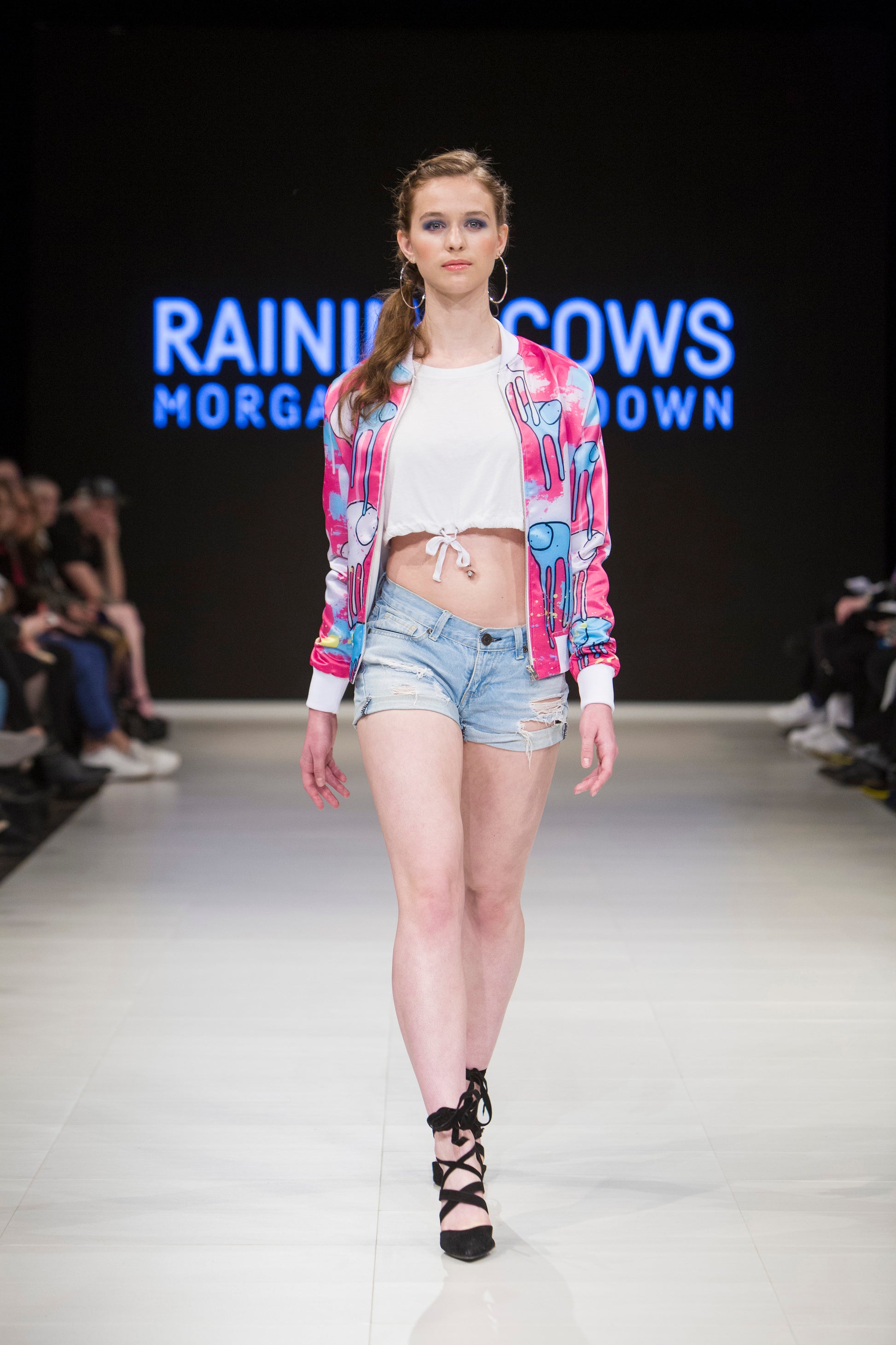 Full-body shot of a model walking the runway wearing a pink and blue multicolored RAINING COWS bomber jacket by Morgan Sheardown, styled with a white crop top, blue denim shorts, black high heels, round neckline, and zipper front.