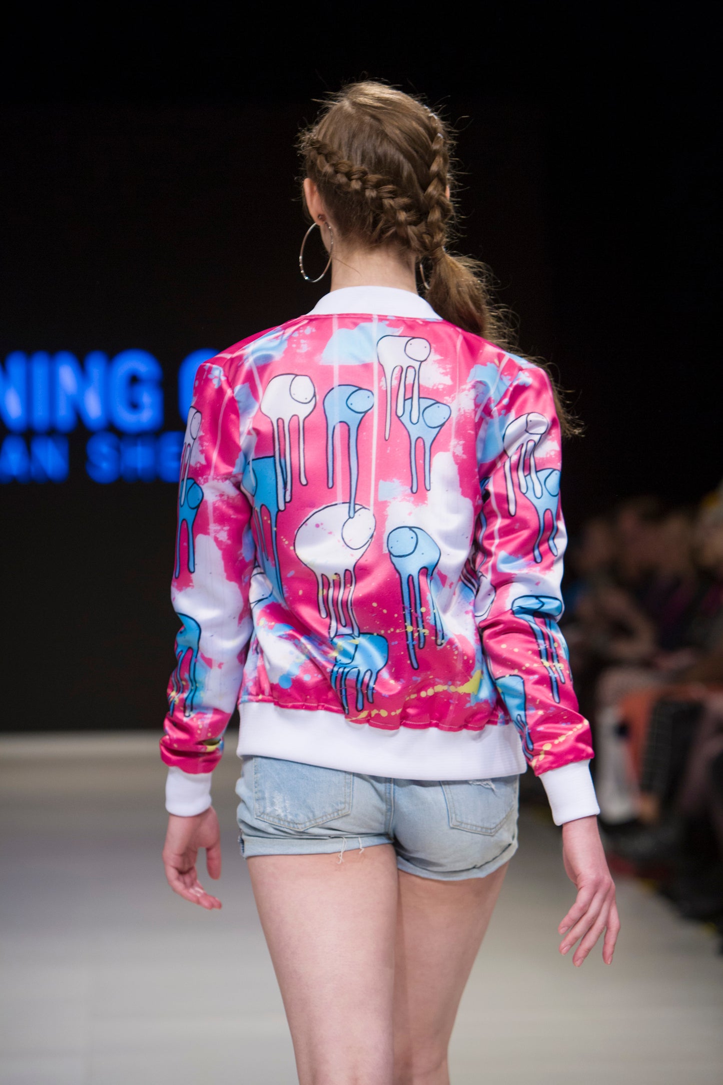Close-up of a model turned around on the runway showing the back of a pink and blue multi coloured RAINING COWS bomber jacket by Morgan Sheardown, paired with blue denim shorts, with seated audience members in the background.