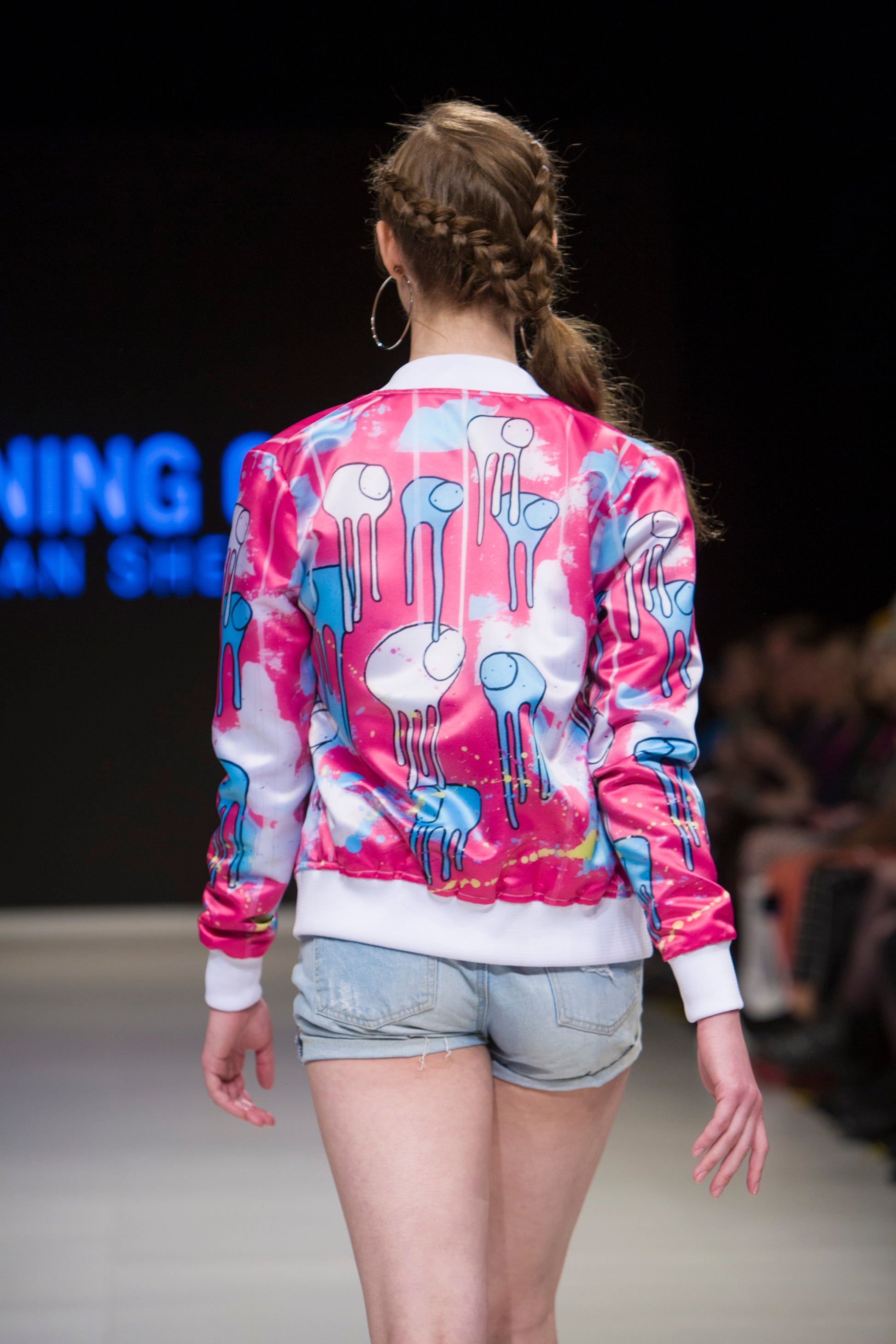 Close-up of a model turned around on the runway showing the back of a pink and blue multi coloured RAINING COWS bomber jacket by Morgan Sheardown, paired with blue denim shorts, with seated audience members in the background.