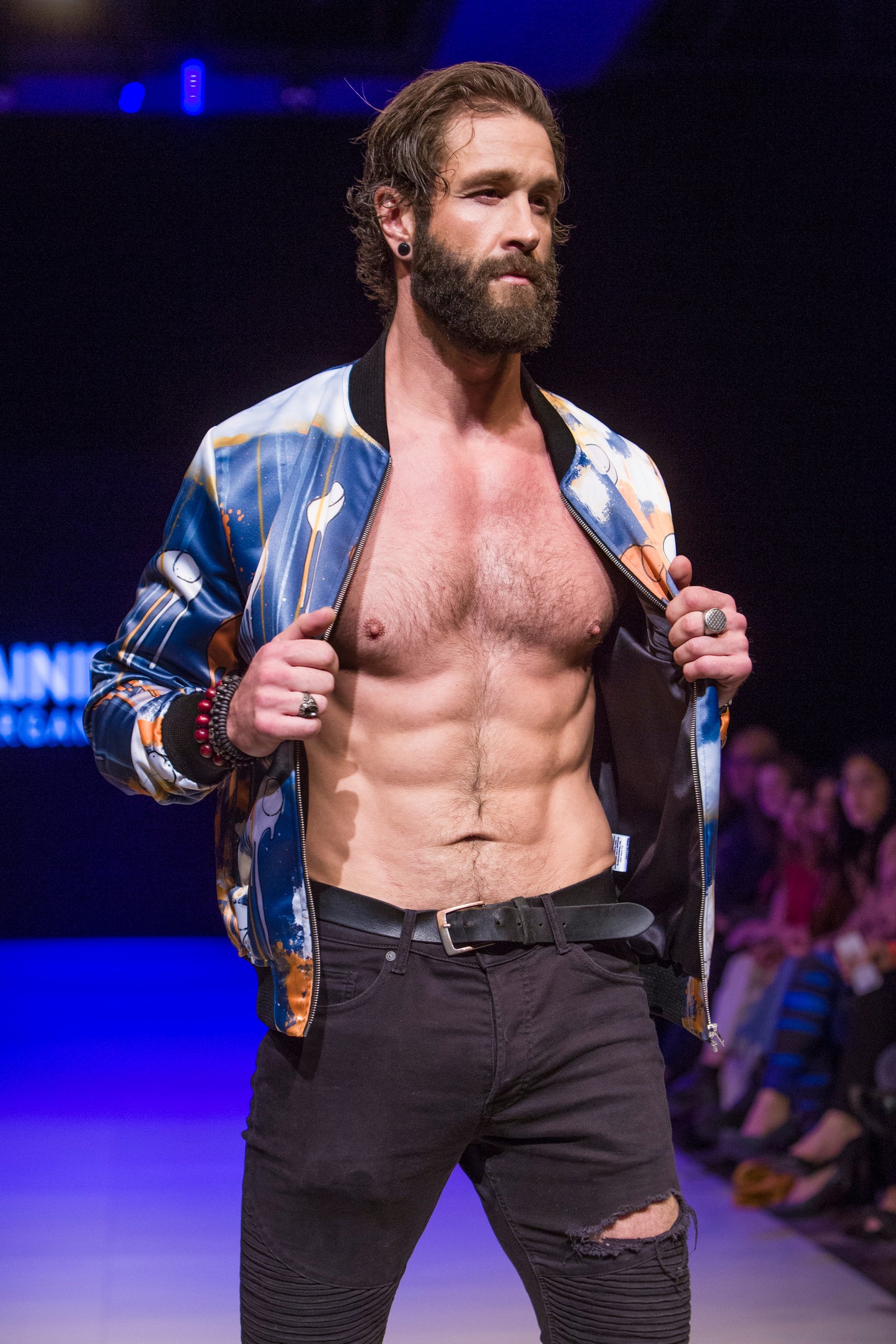 Close-up of a model  on the runway showing the front of a blue and orange multi coloured RAINING COWS bomber jacket by Morgan Sheardown, paired with black pants, with seated audience members in the background.