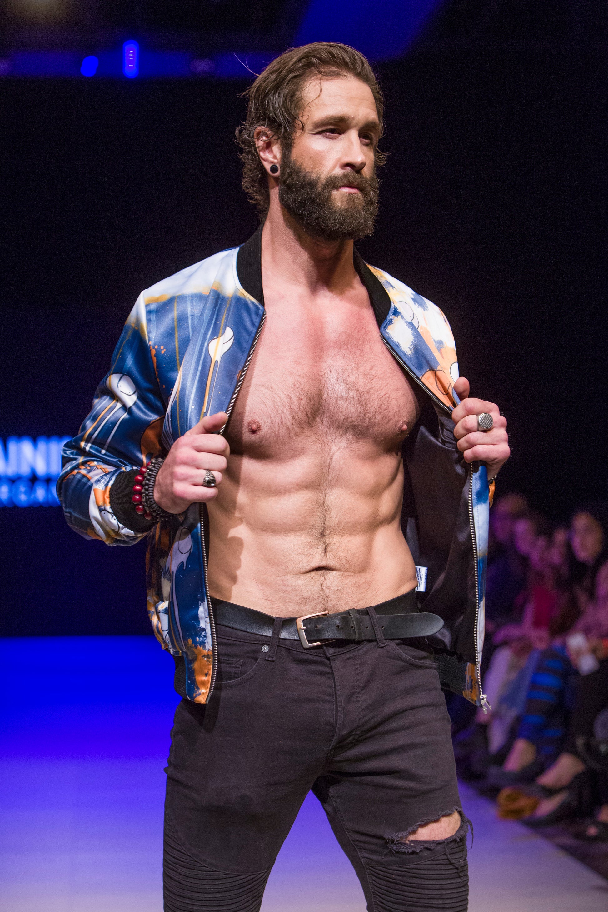 Close-up of a model  on the runway showing the front of a blue and orange multi coloured RAINING COWS bomber jacket by Morgan Sheardown, paired with black pants, with seated audience members in the background.