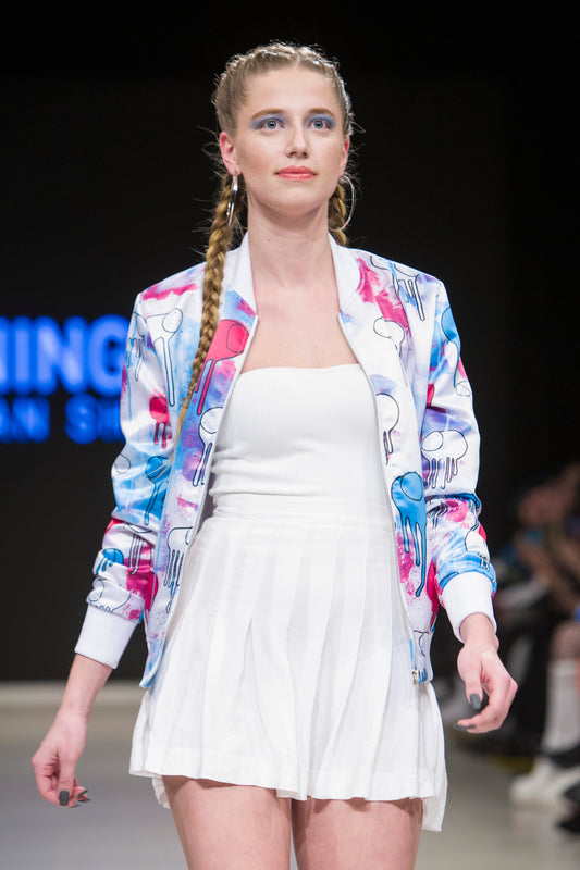 Female model wearing RAINING COWS ‘Cotton Milk’ bomber jacket by Morgan Sheardown, off-white cotton with cow and rain motif, styled with white mini skirt, walking on the runway.