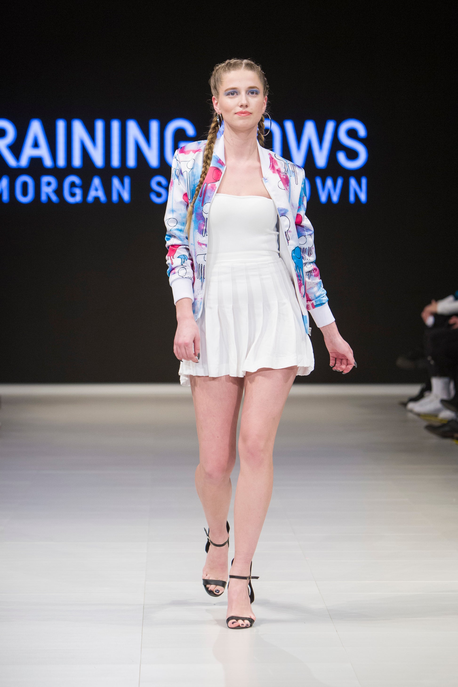 Full-body shot of a model walking the runway wearing a white, blue and pink multi coloured RAINING COWS bomber jacket by Morgan Sheardown, styled with a white crop top, white mini skirt, black high heels.