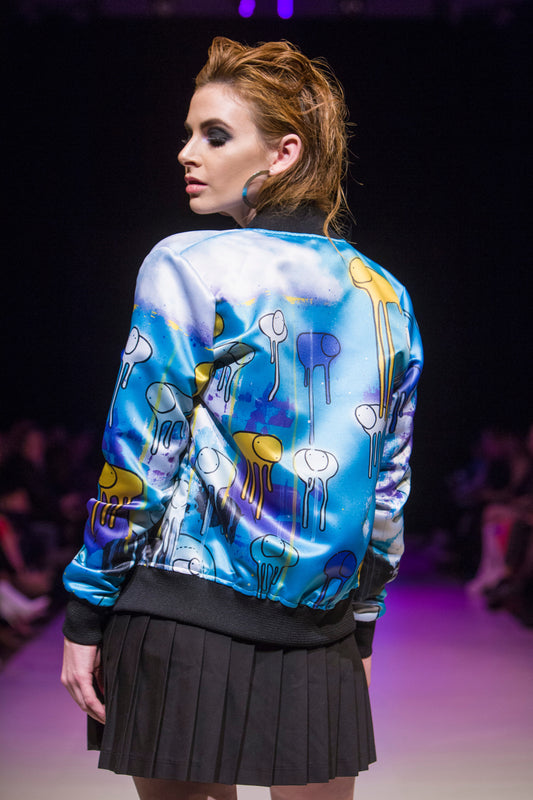 Close-up of a female model turned around on the runway showing the back of a green, purple and yellow multi coloured RAINING COWS bomber jacket by Morgan Sheardown, paired with black mini skirt with seated audience members in the background.