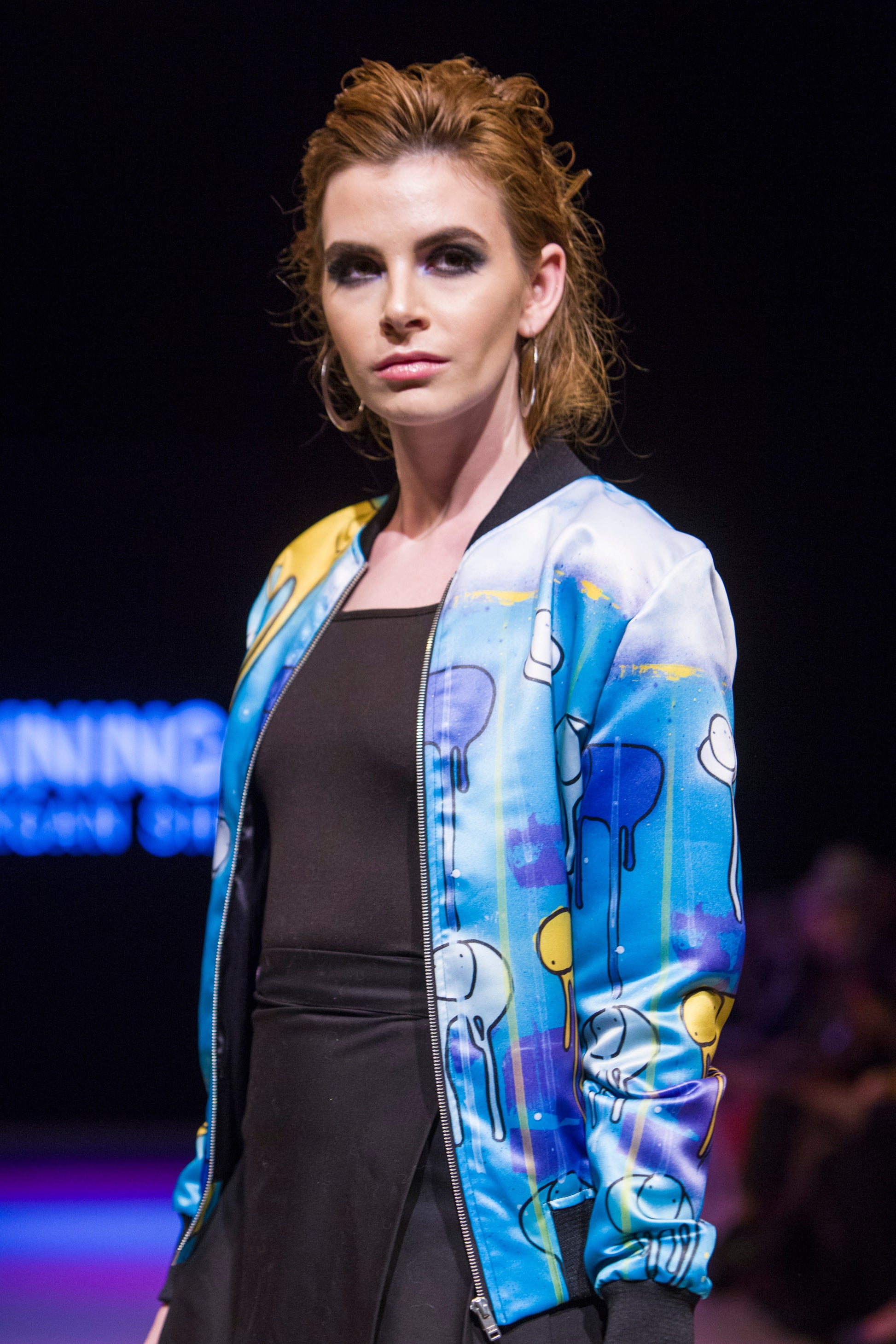 Full-body shot of a model posing on the  runway wearing a green, purple and yellow multi coloured RAINING COWS bomber jacket by Morgan Sheardown, styled with a black top, black skirt, round neckline, and zipper front.