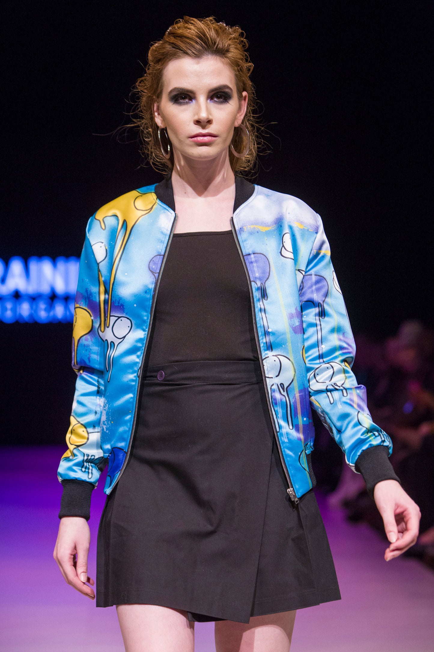 Female model wearing RAINING COWS ‘Guardians of the Sky'  bomber jacket by Morgan Sheardown, green, purple, yellow silk with cow and rain motif, styled with black skirt, posing on the runway.