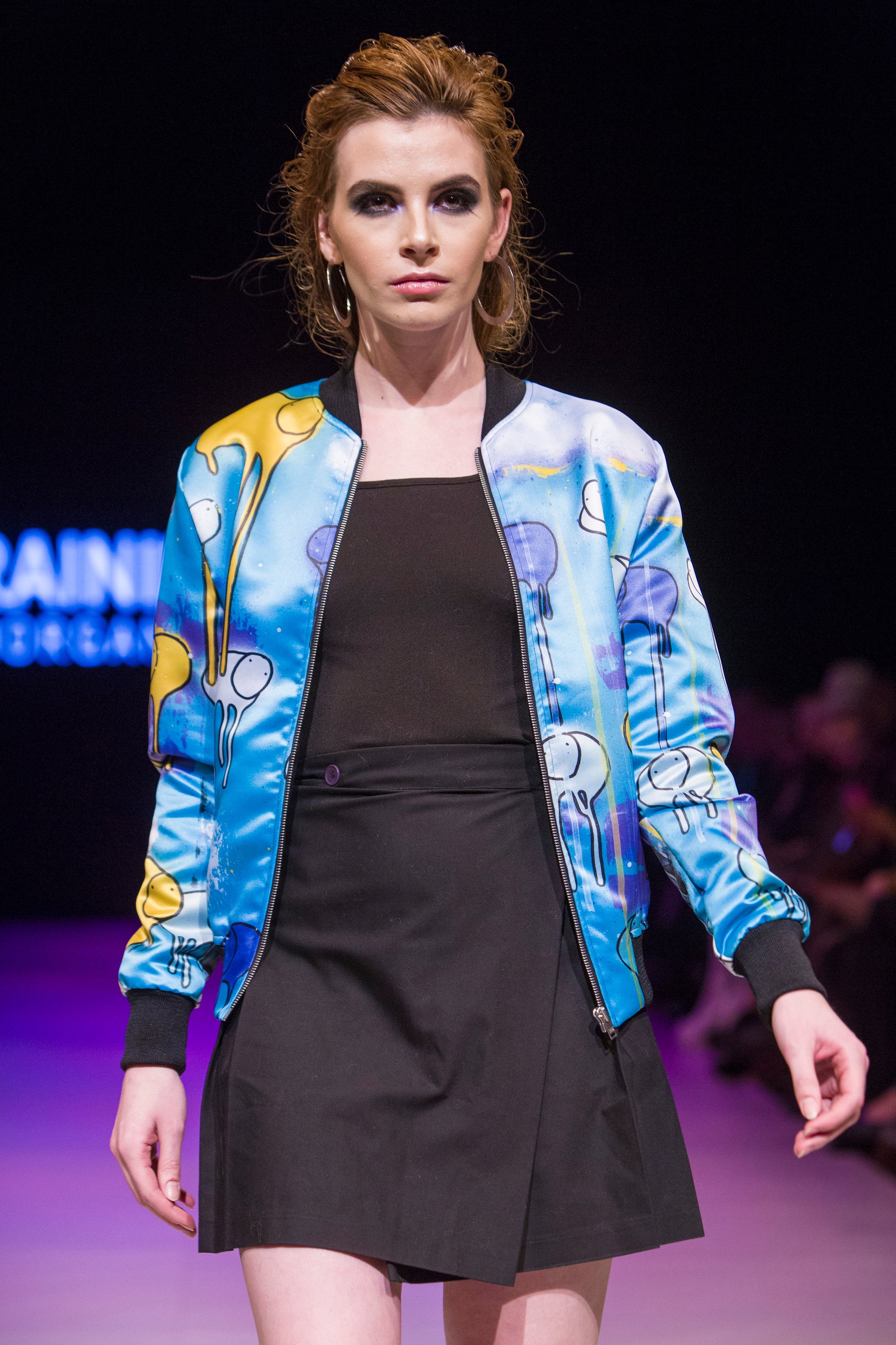 Female model wearing RAINING COWS ‘Guardians of the Sky'  bomber jacket by Morgan Sheardown, green, purple, yellow silk with cow and rain motif, styled with black skirt, posing on the runway.