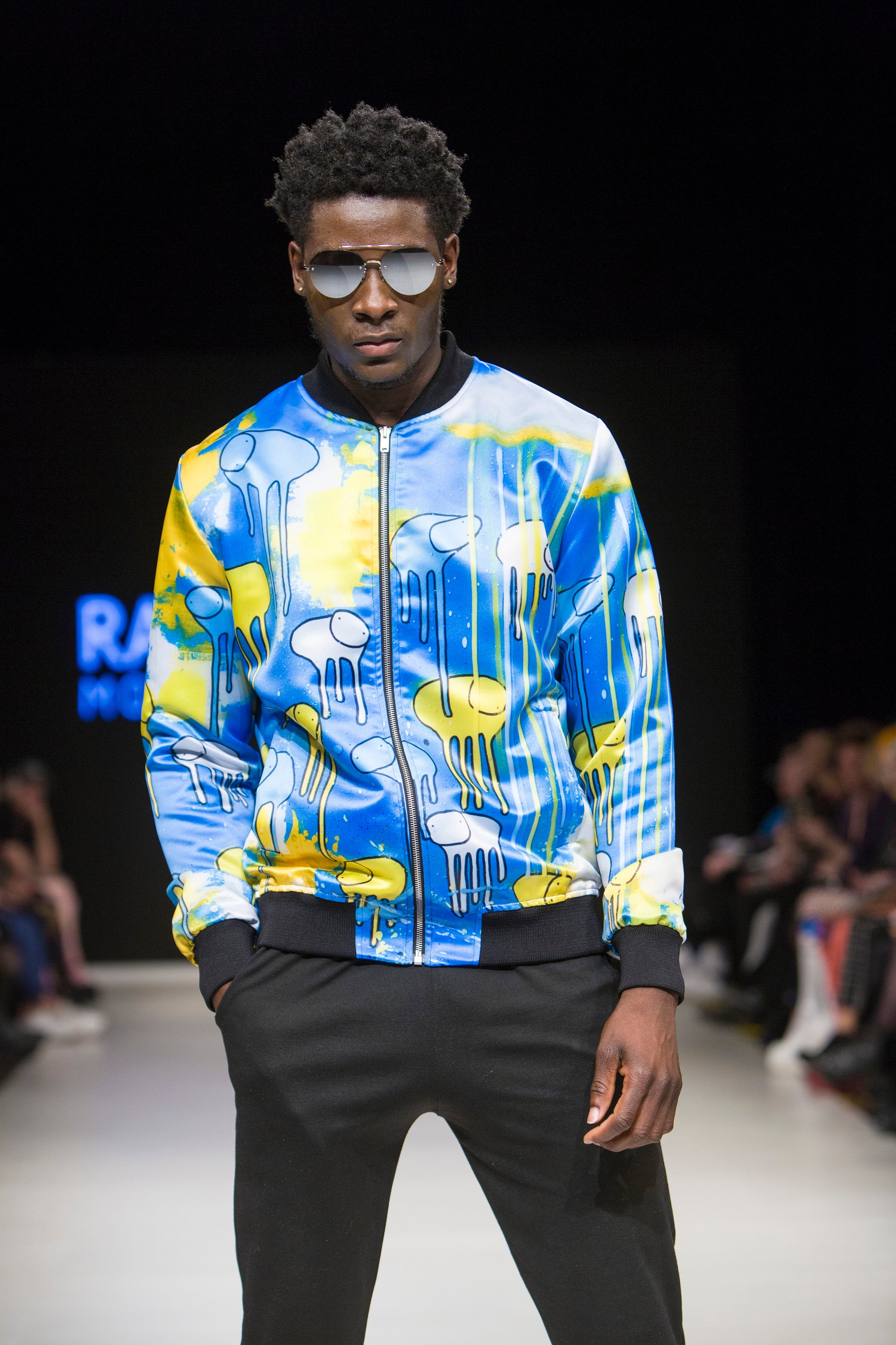 Male model wearing RAINING COWS ‘I See Blue’ bomber jacket by Morgan Sheardown, yellow and blue silk with cow and rain motif, styled with black pants, sunglasses, posing on the runway.