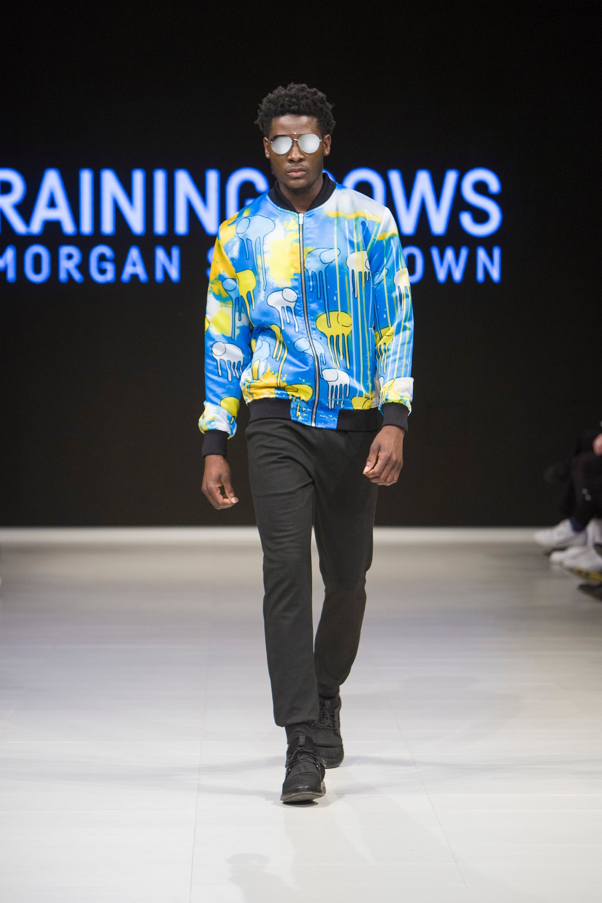 Full-body shot of a model walking the runway wearing a blue and yellow multi coloured RAINING COWS bomber jacket by Morgan Sheardown, styled with black pants, black shoes, round neckline, and zipper front.