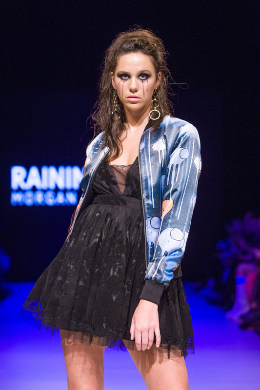Female model wearing RAINING COWS ‘Midnight Symphony' bomber jacket by Morgan Sheardown, blue, purple orange silk with cow and rain motif, styled with black skirt, mascara dripping on her face posing on the runway.