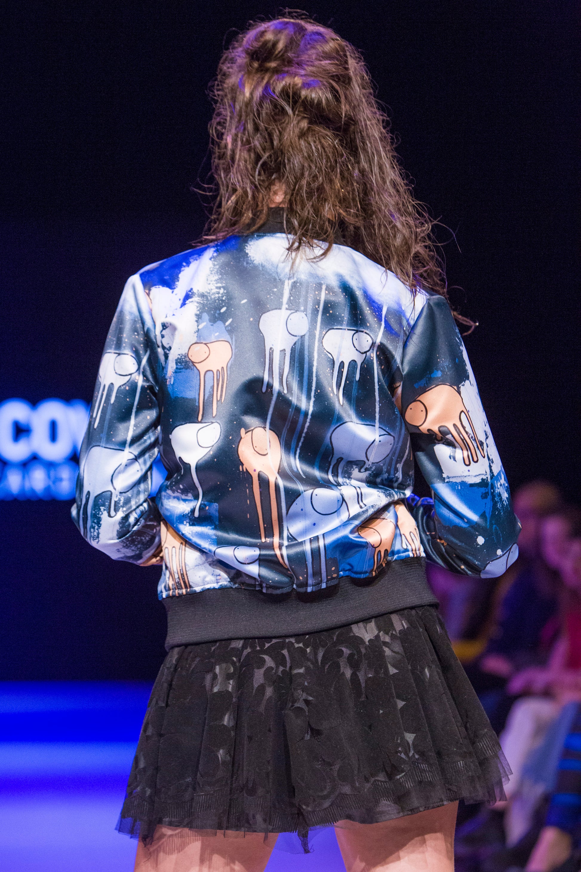Close-up of a model turned around on the runway showing the back of a blue, purple and blue multi coloured RAINING COWS bomber jacket by Morgan Sheardown, paired with a black skirt with seated audience members in the background.