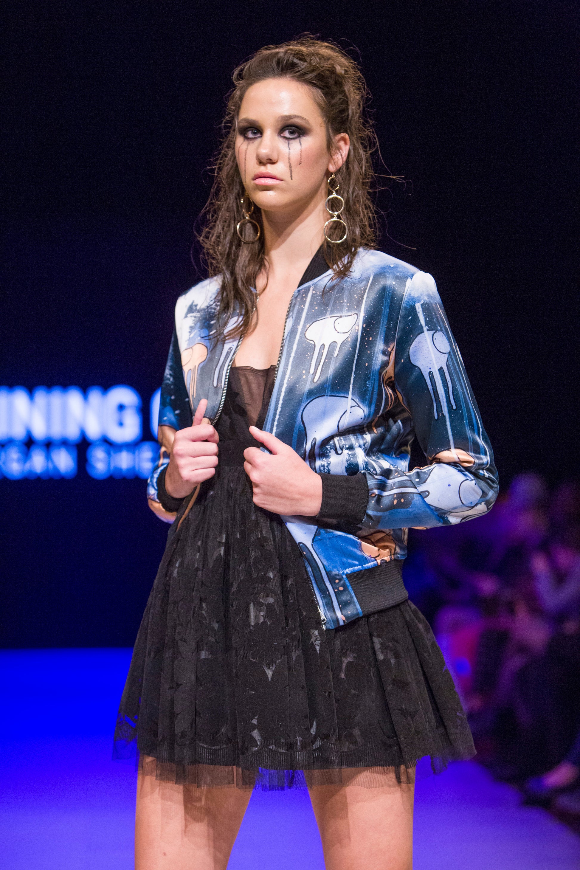 Model posing with mascara running down her face on the runway wearing a blue, purple and orange multi coloured RAINING COWS bomber jacket by Morgan Sheardown, styled with black dress. 
