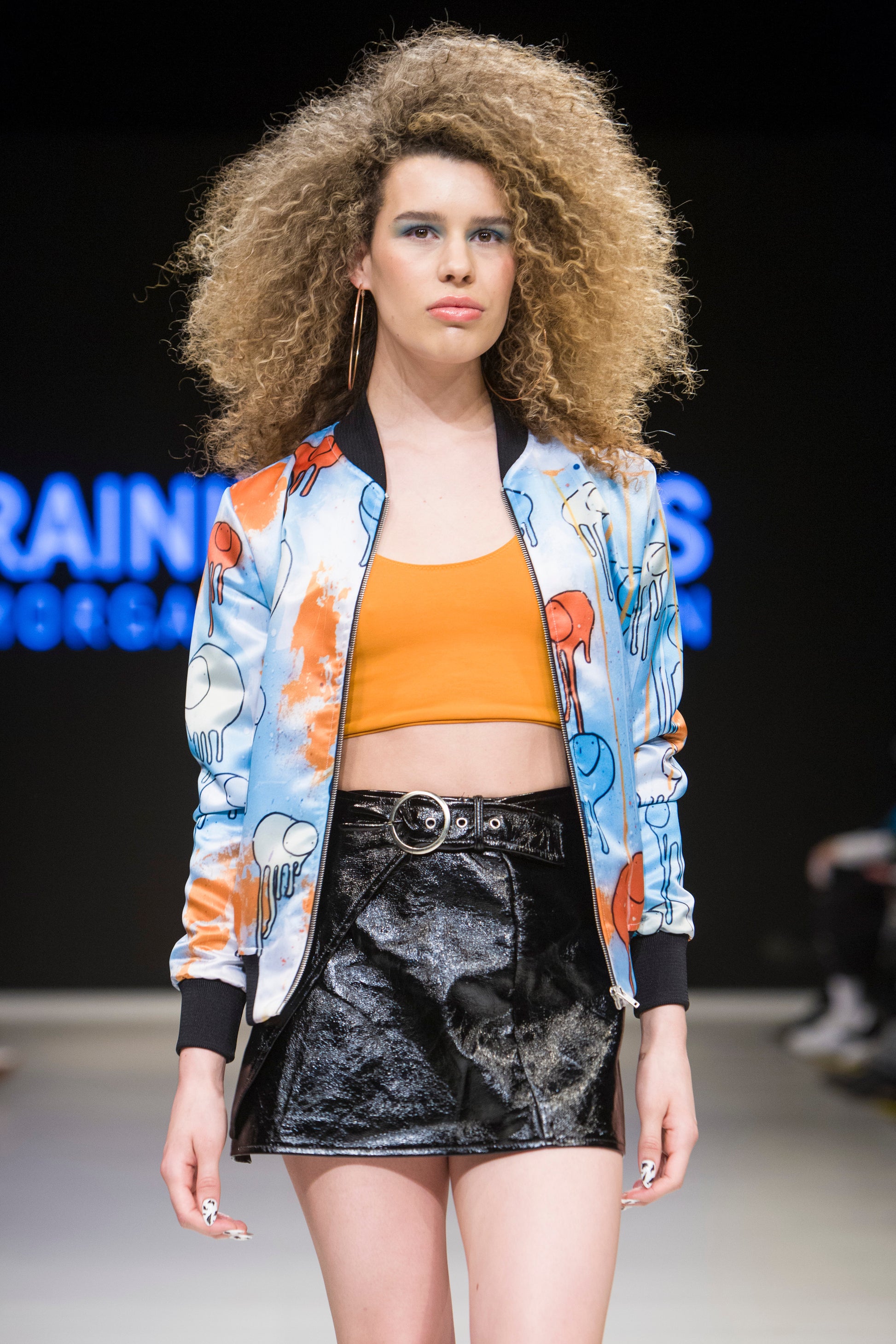 Female model wearing RAINING COWS ‘Powder Blue Sunrise’ bomber jacket by Morgan Sheardown, orange and blue silk with cow and rain motif, styled with orange crop top, black mini skirt, posing on the runway.