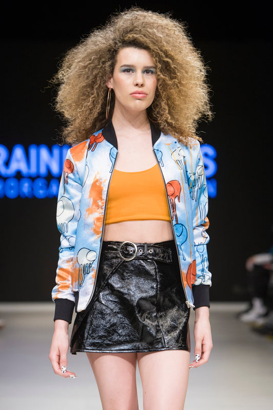 Female model wearing RAINING COWS ‘Powder Blue Sunrise’ bomber jacket by Morgan Sheardown, orange and blue silk with cow and rain motif, styled with orange crop top, black mini skirt, posing on the runway.