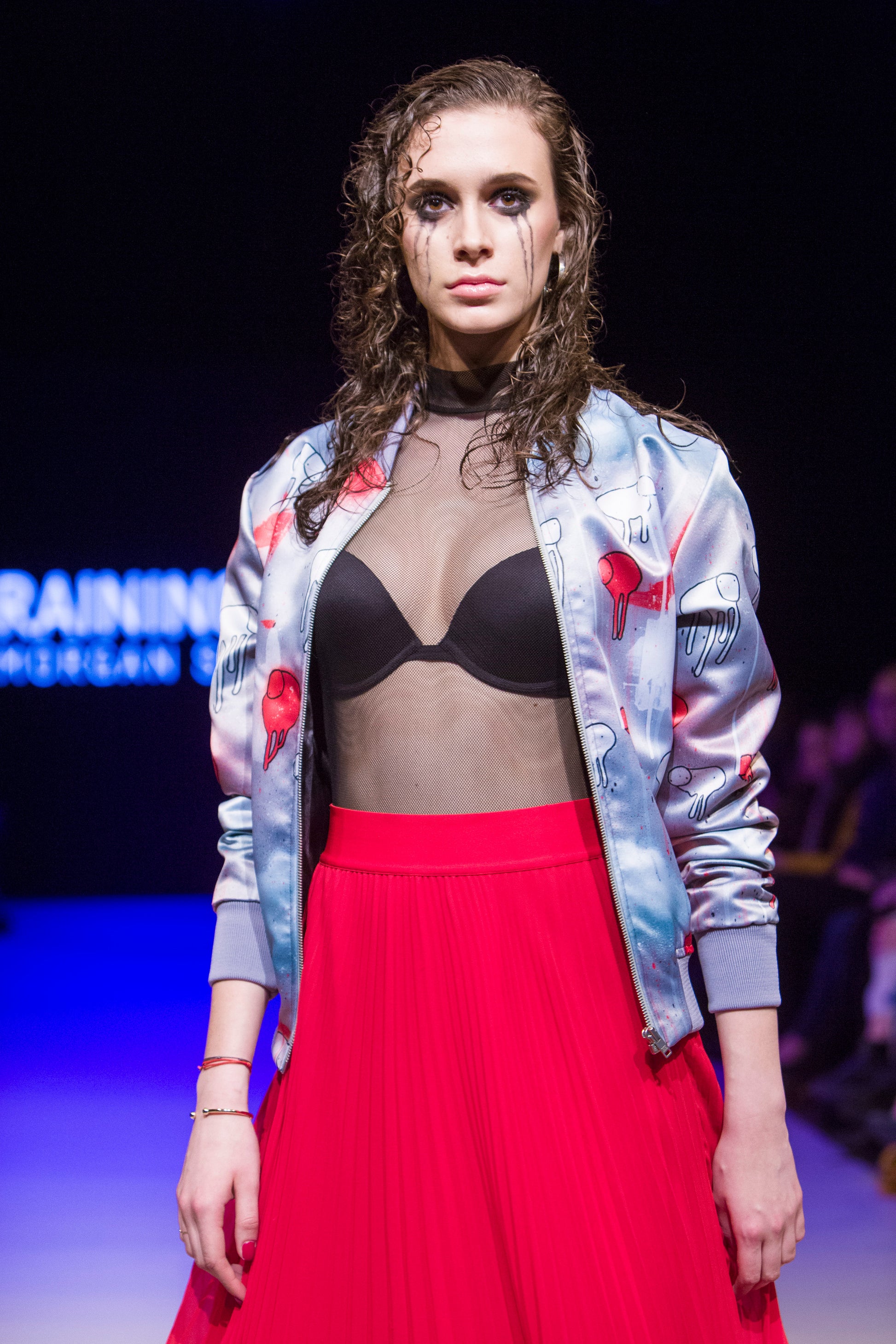 Female model wearing RAINING COWS ‘Red Falls Night' bomber jacket by Morgan Sheardown, red and grey silk with cow and rain motif, styled with black bra, red skirt, mascara dripping on her face posing on the runway.