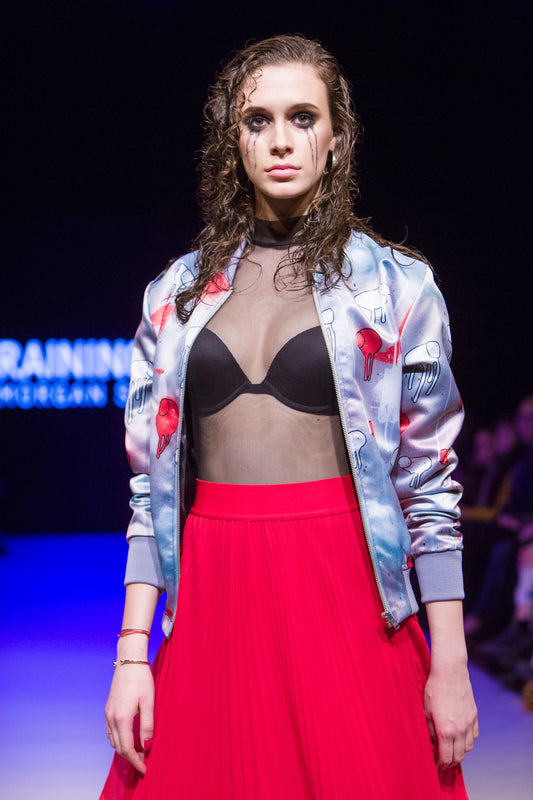 Female model wearing RAINING COWS ‘Red Falls Night' bomber jacket by Morgan Sheardown, red and grey silk with cow and rain motif, styled with black bra, red skirt, mascara dripping on her face posing on the runway.