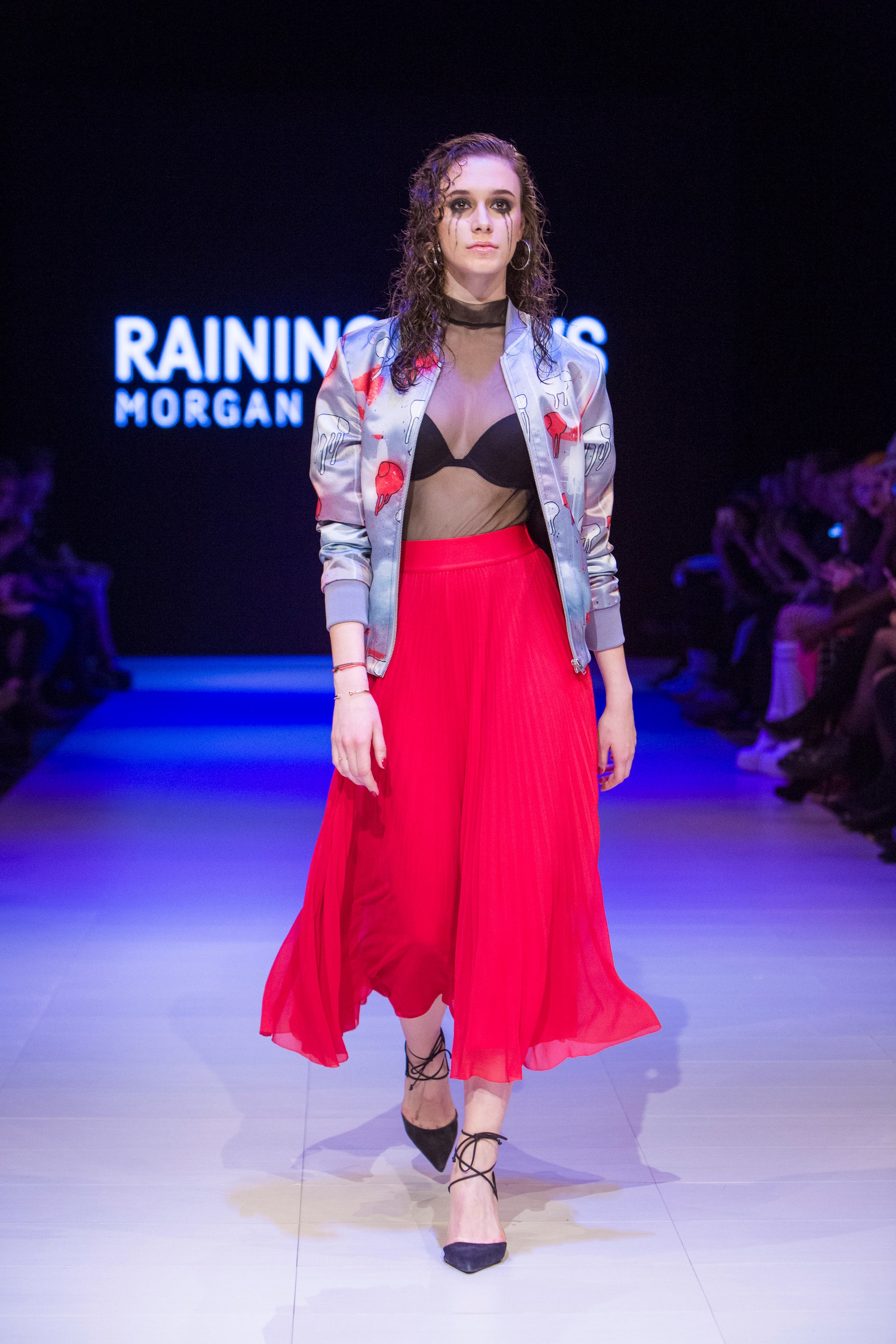 Full-body shot of a model walking the runway wearing a grey and red multi coloured RAINING COWS bomber jacket by Morgan Sheardown, styled with a black bra, red dress, black high heels.