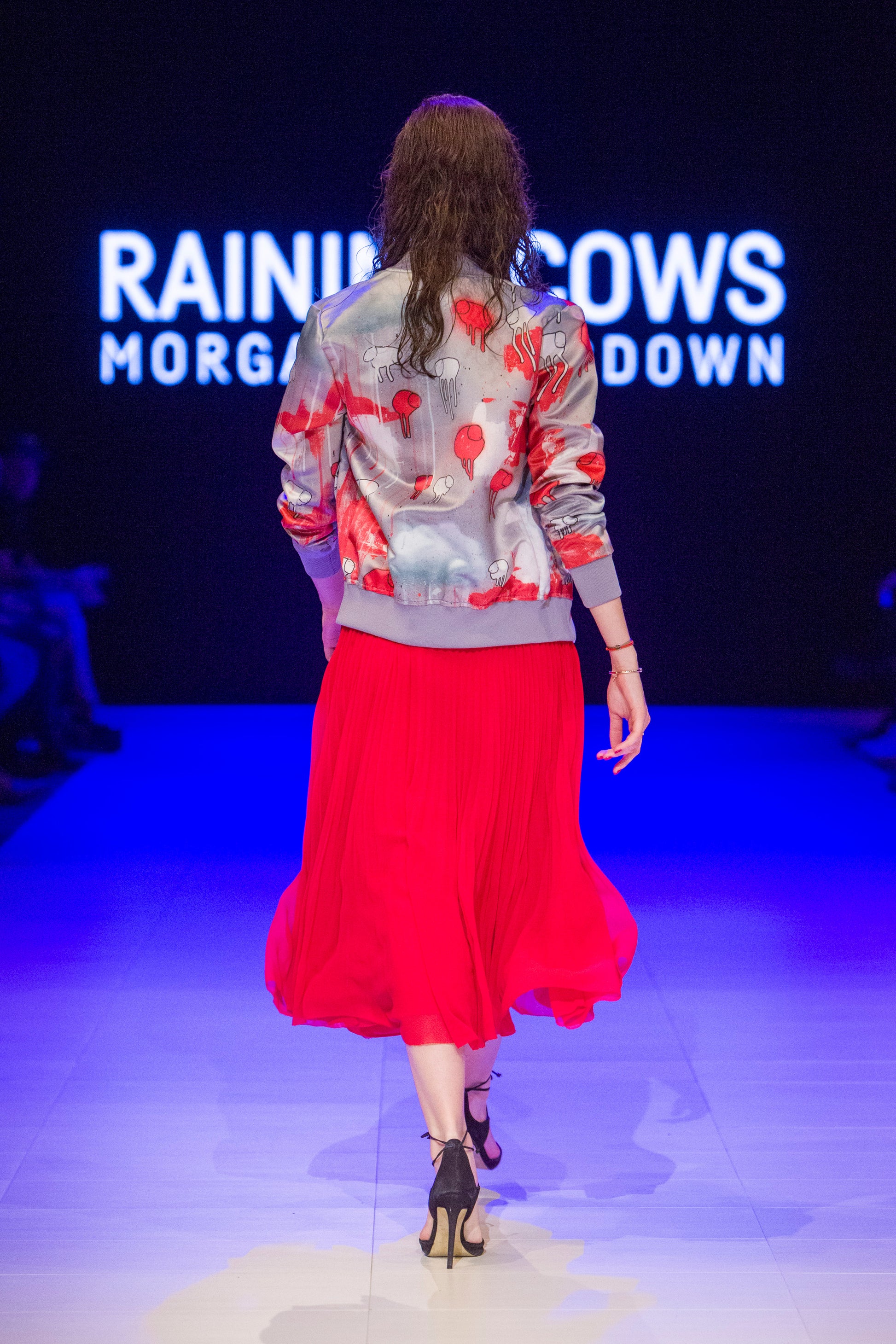 Close-up of a model turned around on the runway showing the back of a grey and red multi coloured RAINING COWS bomber jacket by Morgan Sheardown, paired with red dress and high heals.
