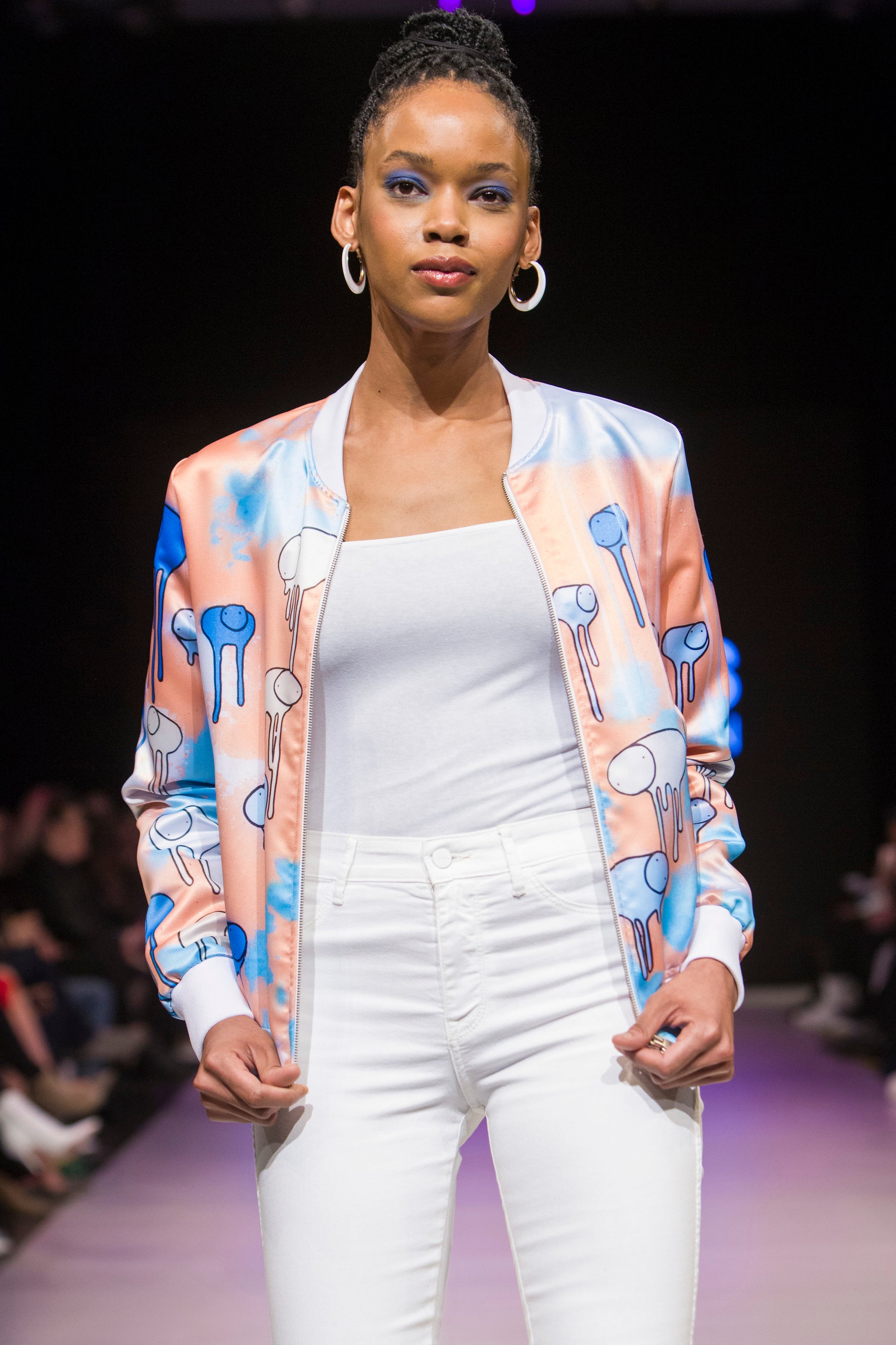 Female model wearing RAINING COWS ‘Salmon Sunset’ bomber jacket by Morgan Sheardown, orange and blue silk with cow and rain motif, styled with white top, white pants, posing on the runway.