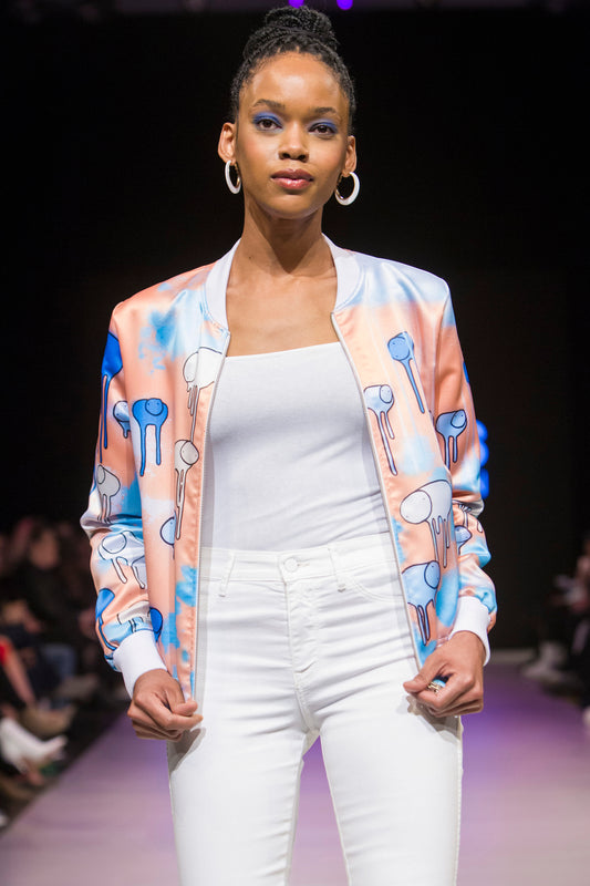 Female model wearing RAINING COWS ‘Salmon Sunset’ bomber jacket by Morgan Sheardown, orange and blue silk with cow and rain motif, styled with white top, white pants, posing on the runway.