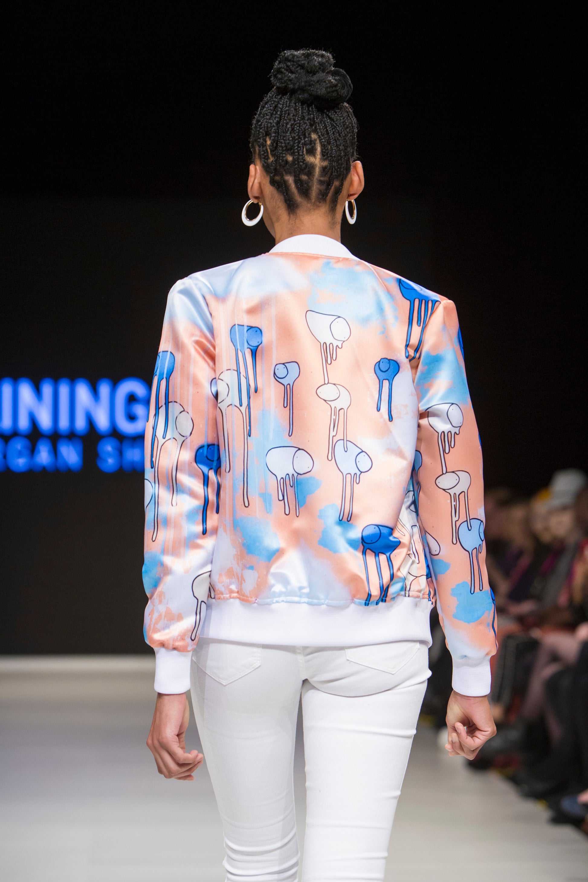 Close-up of a model turned around on the runway showing the back of a orange and blue multi coloured RAINING COWS bomber jacket by Morgan Sheardown, paired with white pants, with seated audience members in the background.