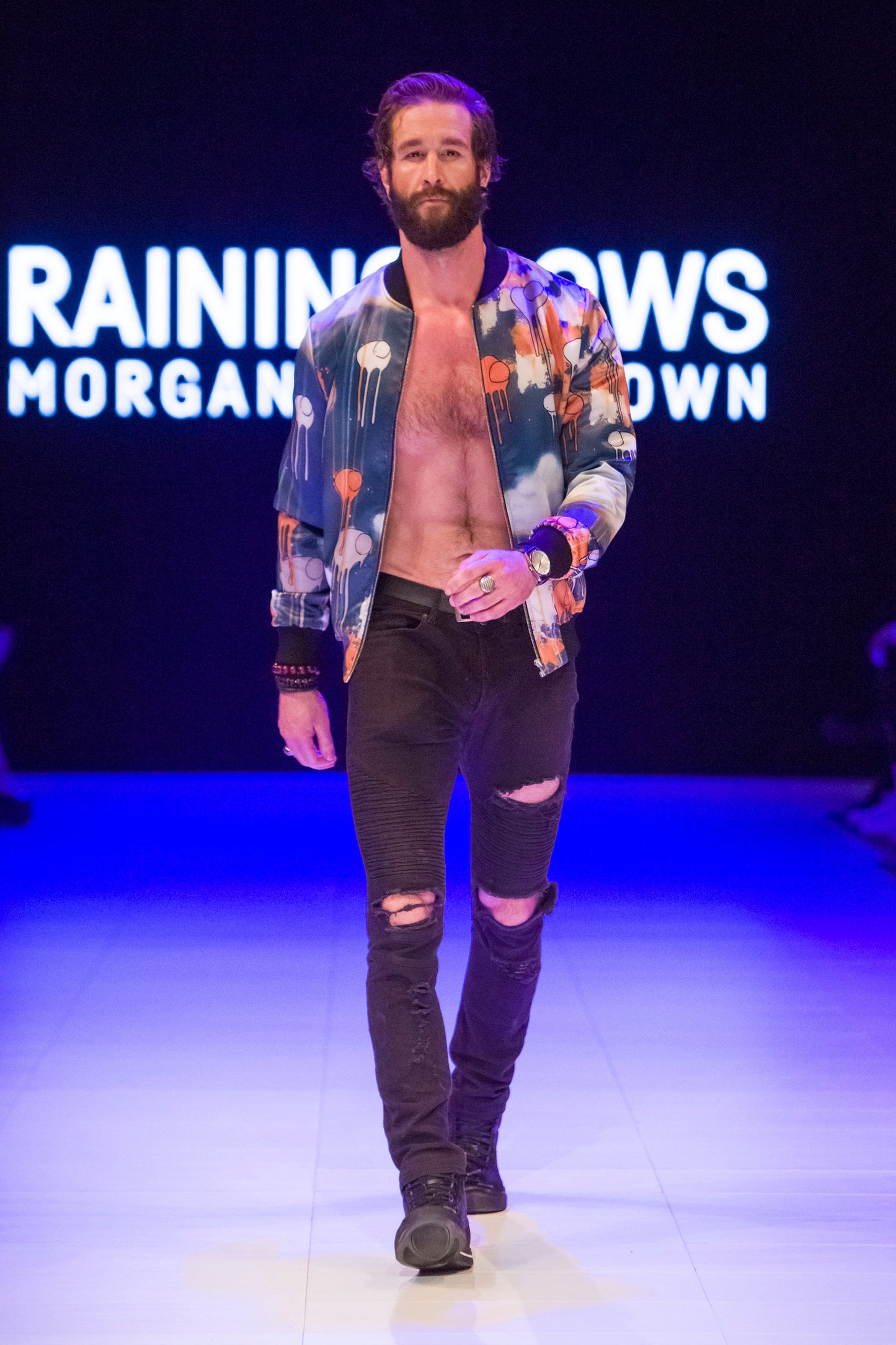 Full-body shot of a model walking the runway wearing a blue and orange multi coloured RAINING COWS bomber jacket by Morgan Sheardown, styled with black pants, black shoes, round neckline, and zipper open in the front.