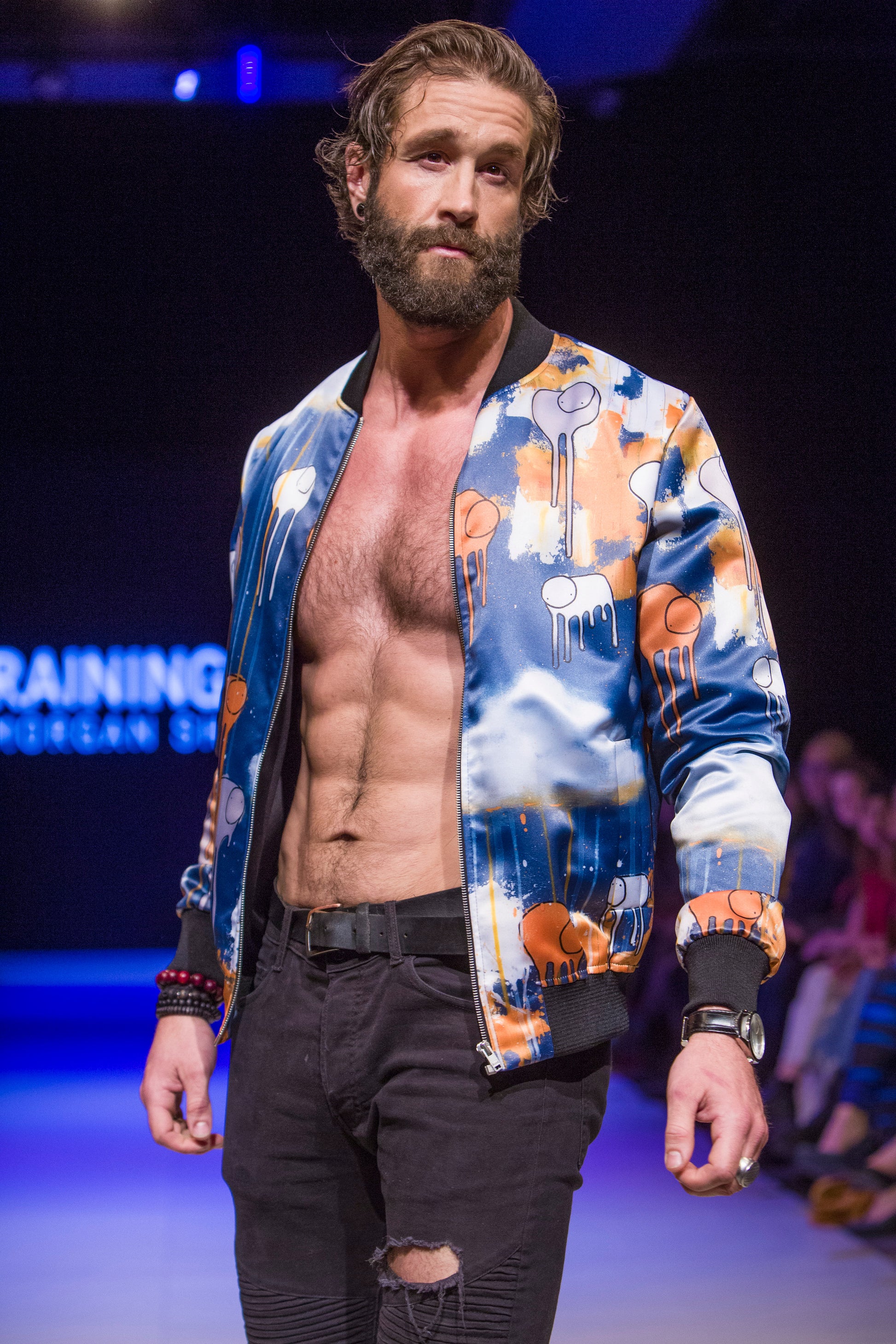 Male model wearing RAINING COWS ‘Tears of the Sun’ bomber jacket by Morgan Sheardown, orange and blue silk with cow and rain motif, styled with black pants, posing on the runway.